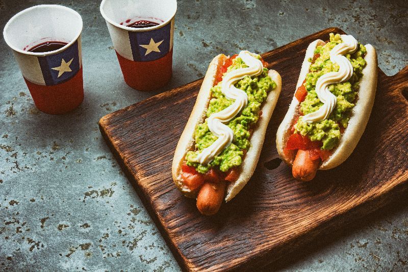 Chilean Completo Italiano. Hot dog sandwiches with tomato, avocado and mayonnaise served on wooden board with drink in paper cup . Top view. Independence Day concept.